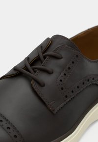 Chaussure en cuir marron avec détails perforés, bout arrondi et lacets plats. Elle présente une semelle en caoutchouc blanc et une finition lisse.