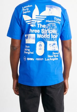T-shirt bleu avec texte blanc et graphiques listant des villes mondiales et "The three Stripes World tour" au dos.