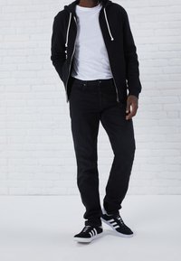Sudadera negra con cremallera y cordones blancos, camiseta blanca, jeans oscuros y zapatillas negras con rayas blancas y negras.