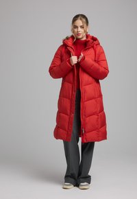 Rode pufferjas met een capuchon, voorzien van een ritssluiting, gestepelde textuur en zijzakken. Draag het over een rode trui en losse grijze broek.