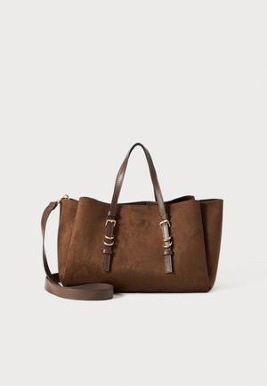 Sac cabas en suède marron avec poignées réglables marron foncé et bandoulière amovible sur un fond clair uni.