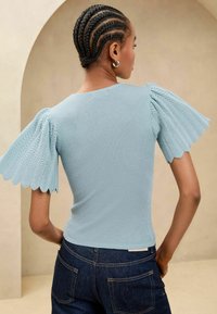 Top rib de culoare bleu deschis, cu mâneci texturate și cu margini ondulate. Design ajustat cu decolteu rotund, asortat cu blugi de denim închis.