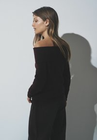Burgundy off-the-shoulder sweater with ribbed texture, long sleeves, and a relaxed fit, paired with dark trousers. Soft fabric detail visible.