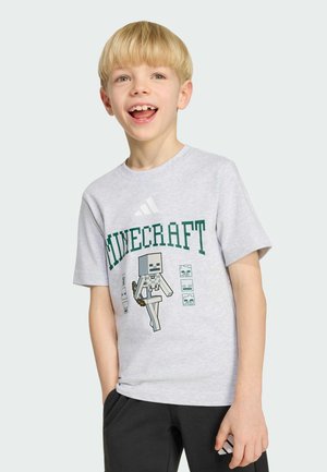 Niño rubio sonriendo con la mano derecha en el bolsillo, vistiendo una camiseta gris de Minecraft y pantalones negros sobre un fondo claro.