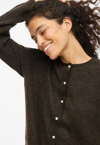 Smilende kvinne med krøllete hår løfter armen, iført en mørk brun cardigan med perleknapper mot en lys bakgrunn.