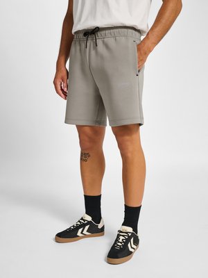 Mand iført grå snor-shorts, sorte sokker og sorte sneakers med hvide detaljer, stående mod en hvid baggrund.