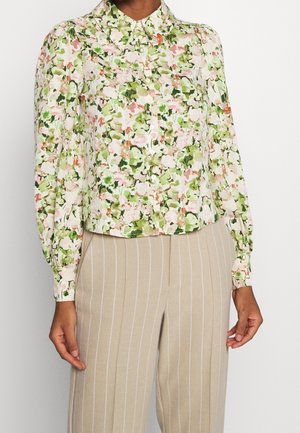 Kvinde iført bluse med lange ærmer, blomsterprint i grønne, pink og hvide farver, kombineret med beige bukser med fine striber.