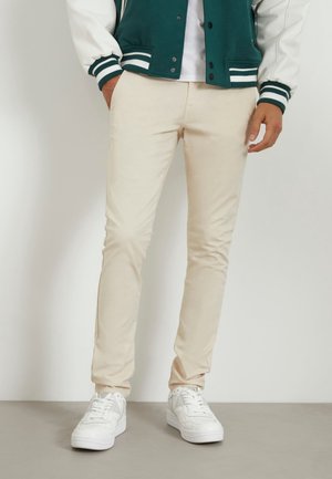 Mężczyzna ubrany w beżowe, dopasowane spodnie, białe trampki, białą koszulkę oraz zieloną kurtkę typu varsity z białymi rękawami i paskowanymi mankietami.