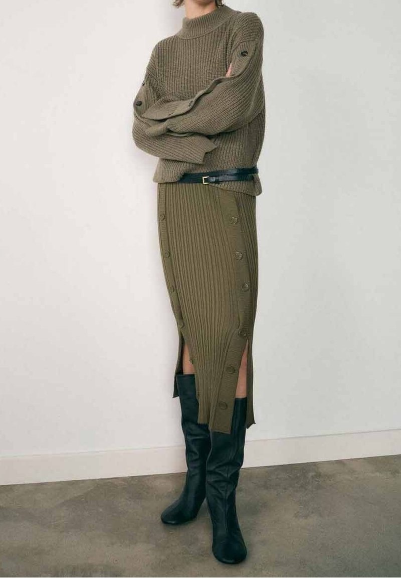 Personne portant un pull en maille côtelée vert olive et une jupe boutonnée assortie avec une ceinture noire et des bottes noires montantes, debout contre un mur blanc.