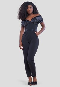 Donna con capelli ricci che indossa una tuta nera off-shoulder con corpino in satin arricciato e tacchi aperti neri con dettagli a fiocco.