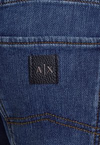 Blauer Denim-Stoff mit einem strukturierten schwarzen Lederpatch. Auf dem Patch sind die Buchstaben "A|X" in metallisch Silber zu sehen. Kontrastnähte sind sichtbar.