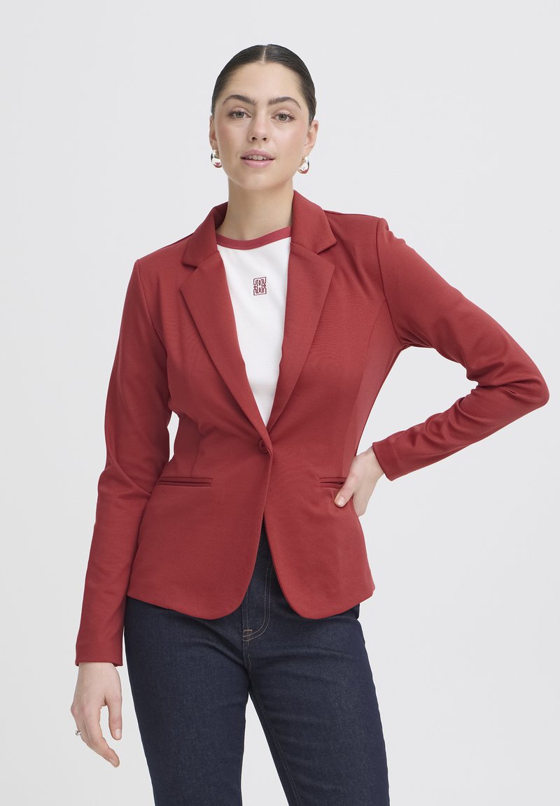 Kvinde iført en tætsiddende rød blazer over en hvid skjorte med logo, kombineret med mørkeblå jeans, poserer med den ene hånd på hoften mod en ensfarvet baggrund.