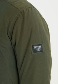 Oliivinvihreä takki, jossa on teksturoitu kangas, kirjailtu malli ja hihassa brändäysyksityiskohtia sisältävä päällyste.