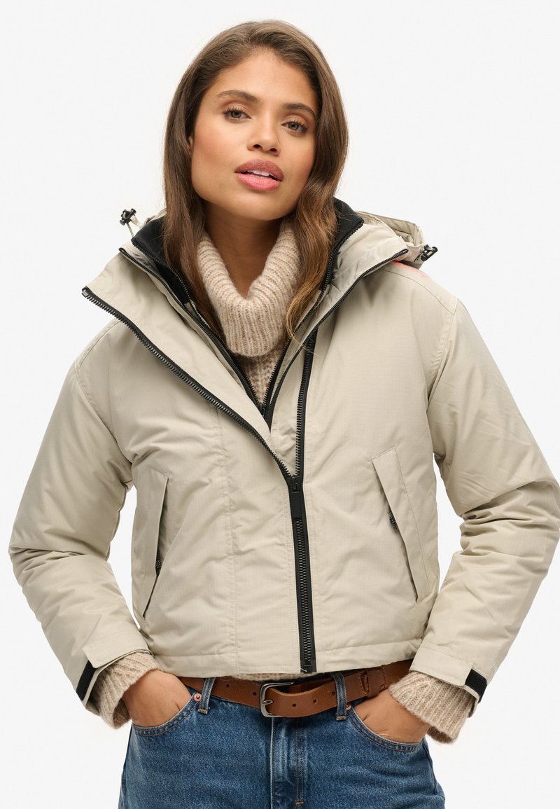Beige cropped jack met een zwarte rits, capuchon en zijzakken. Draagt over een chunky gebreide beige trui, gecombineerd met blauwe jeans.