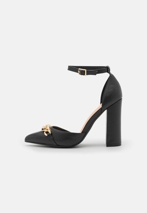 Czarny skórzany but na wysokim obcasie z ostrym noskiem, paskiem wokół kostki i złotym akcentem na przodzie. Obcas typu słupek zapewnia stabilność.