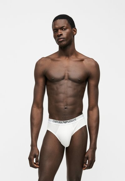 Αθλητικός άνδρας με σκουρόχρωμο δέρμα, ντυμένος με λευκά εσώρουχα Emporio Armani, στέκεται μπροστά σε απλό ανοιχτόχρωμο φόντο με ουδέτερη έκφραση.