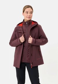 Donkerrode waterdichte jas met capuchon, voorzien van een rode voering aan de binnenkant, een ritssluiting aan de voorkant en meerdere functionele zakken. Zwarte broek eronder gedragen.