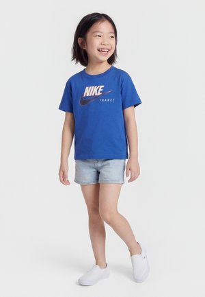 Smilende barn iført en blå Nike France t-skjorte, lyse denimbukser og hvite slip-on sko, stående med én fot lett løftet.