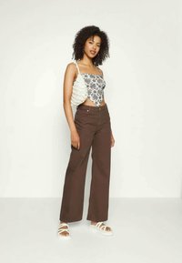 Jovem mulher com cabelo encaracolado, vestindo um top de flores sem mangas, calças largas de cintura alta castanhas e sandálias brancas com tiras, em pé.