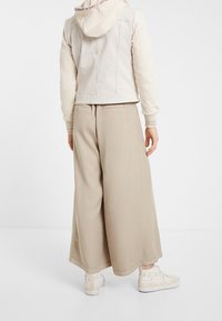 Persona que lleva pantalones de pierna ancha beige, chaqueta de denim blanca roto con capucha y zapatillas color crema con detalles de encaje floral, de espaldas.