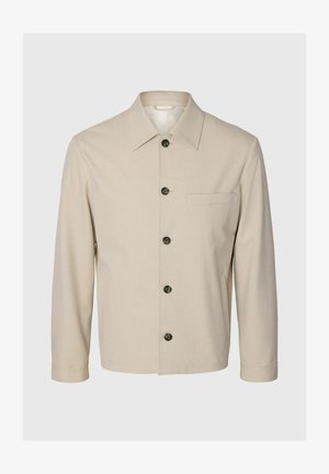 Bézs hosszú ujjú dzseki hegyes gallérral, elülső gombokkal, egy mellkasi díszzsebbel és két oldalsó zsebbel, egyszínű háttéren bemutatva.
