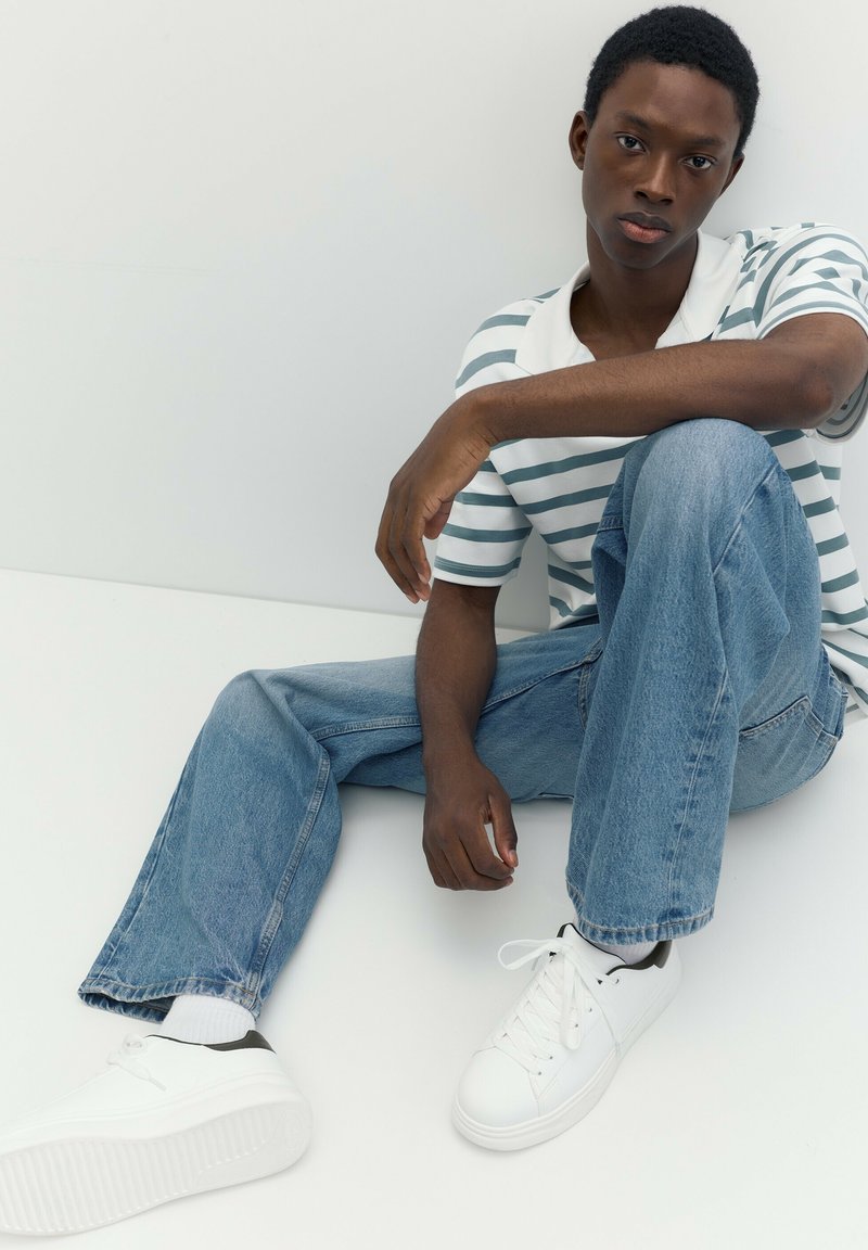 Jeune homme aux cheveux courts portant une chemise rayée, un jean bleu et des baskets blanches, assis par terre contre un mur blanc uni.