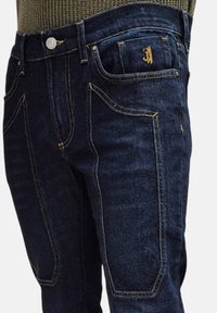 Jeans in denim blu scuro con cuciture gialle a contrasto, un bottone argentato e un design unico delle tasche. Il tessuto ha una texture liscia e una vestibilità strutturata.