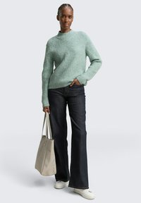 Mintgrüner Ripp-Pullover mit hohem Kragen, kombiniert mit dunklen Schlagjeans und einer hellbeigen Tasche, die aus strukturiertem Material besteht und breite Manschetten hat.