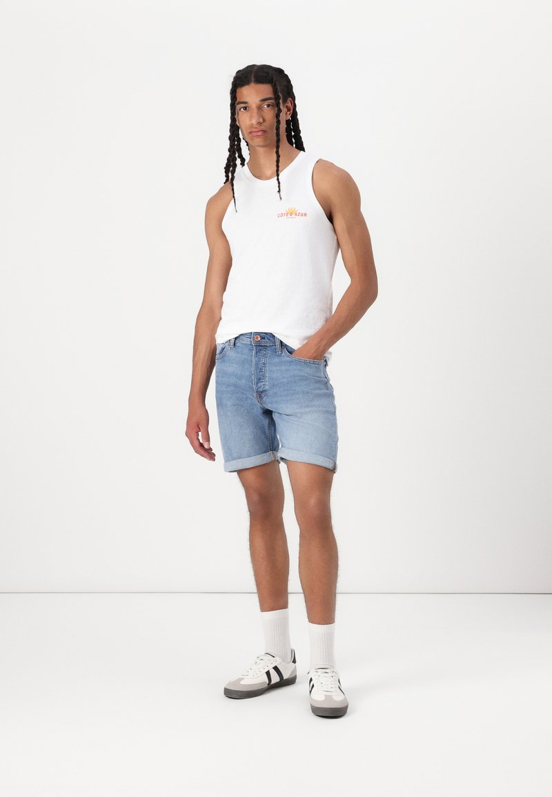 Débardeur blanc sans manches avec impression sur le devant, short en denim bleu clair, chaussettes blanches et chaussures avec accents noir et gris. Design simple, coupe décontractée.