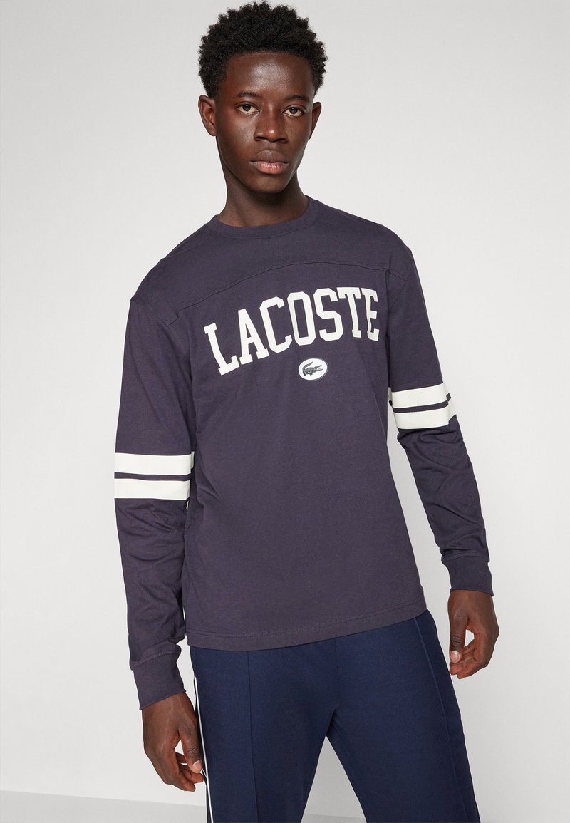 Marineblauwe long-sleeve t-shirt met witte "LACOSTE" tekst en gestreepte mouwaccenten. Gemaakt van zacht katoen met een ronde halslijn.