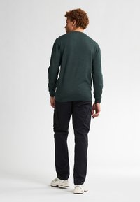 Suéter verde oscuro con cuello redondo y mangas largas, combinado con pantalones cargo negros que tienen bolsillos laterales. Zapatillas blancas completan el look.