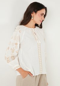 Blusa bianca con ricami floreali beige, scollatura tonda e dettaglio con coulisse. Maniche lunghe con polsini elasticizzati. Tessuto leggero.
