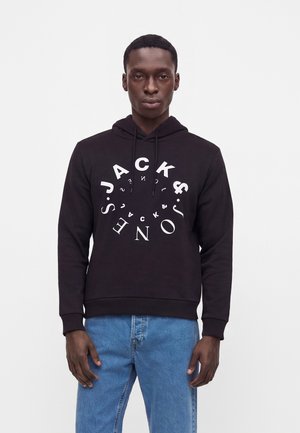Homme portant un sweat à capuche noir avec un texte blanc circulaire et un jean bleu, debout devant un fond blanc uni.