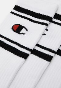 Calcetines de algodón blancos con textura acanalada, que presentan dos rayas negras y un logotipo negro y rojo en un lado.