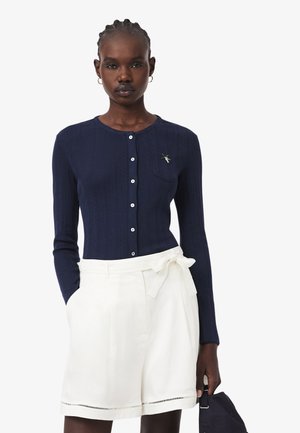 Vrouw draagt een marineblauwe, lange mouwen gebreide cardigan met knopen en een broche, gecombineerd met witte shorts met hoge taille en een ceintuur, en houdt een navy tas vast.