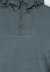 Giacca leggera grigia con cappuccio, chiusura con zip e tasca sul petto. Caratterizzata da una texture liscia e un design minimalista.