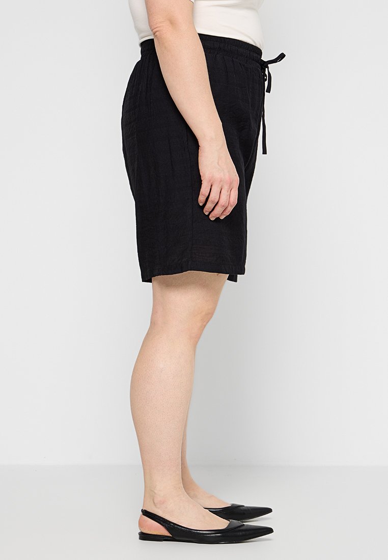Schwarze Leinen-Shorts mit strukturierter Oberfläche, ausgestattet mit einem elastischen Bund und einer Kordel. Getragen mit schwarzen spitzen Ballerinas.