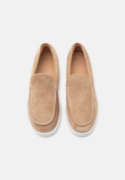 Ζευγάρι μπεζ μοκασίνια από σουέντ με λευκές σόλες, λεπτομερής ραφή στην κορυφή, φωτογραφημένα από πάνω σε λευκό φόντο.