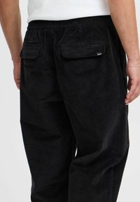 Black corduroy pants with a relaxed fit, featuring two back pockets and an elastic waistband. Surface has a ribbed texture.