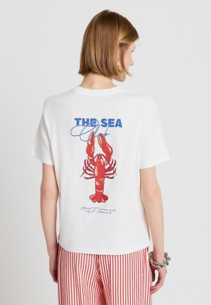 Donna con capelli corti che indossa una maglietta bianca con un'aragosta rossa e la scritta "THE SEA", abbinata a pantaloni a righe rosse e bianche.