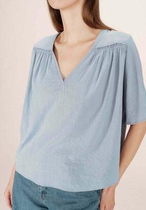 Blouse bleu clair ample avec un col en V, des manches courtes et de subtils plis verticaux le long des épaules, fabriquée en tissu texturé.