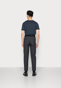 En man som bär en mörkblå t-shirt, grå rutig byxa med ett svart bälte, svarta strumpor och svarta läderskor, står med ryggen mot kameran.