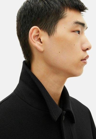 Giovane con capelli neri corti che indossa un cappotto nero con colletto, mostrato di profilo contro uno sfondo bianco semplice.