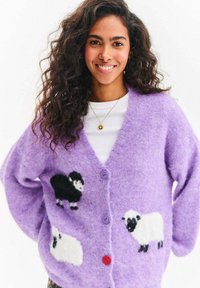 Młoda kobieta o kręconych włosach uśmiecha się, nosząc lawendowy kardigan z wzorem czarnych i białych owiec oraz kolorowymi guzikami, na białej koszuli.