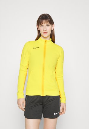 Gelbe sportliche Zip-Jacke mit hohem Kragen, schwarzem Nike-Logo, figurbetontem Design und langen Ärmeln. Kombiniert mit schwarzen Shorts mit weißem Logo.