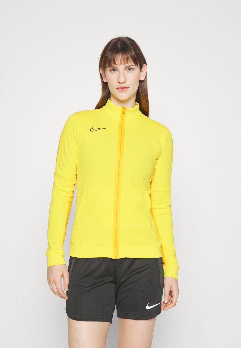 Yellow athletic zip-up jacket with a high collar, black Nike logo, fitted design, and long sleeves. Paired with black shorts featuring a white logo.