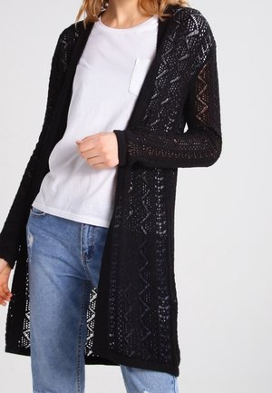 Person iført en hvid t-shirt, lyseblå jeans og en lang sort åbenstrikket cardigan med geometrisk mønster.