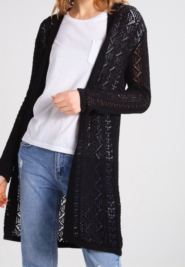 Personne portant un t-shirt blanc, un jean bleu clair et un long cardigan noir en maille ajourée avec un motif géométrique.