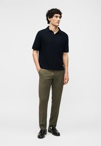 Άνδρας με σγουρά μαλλιά, φορώντας μαύρο πόλο με κοντά μανίκια, ελαιδί παντελόνι και μαύρα παπούτσια, στέκεται με το ένα χέρι στην τσέπη.