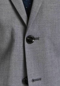 Grå blazer med tekstureret stof, to sorte knapper og en diskret klapkrave. Indersiden har en marineblå foring med små mønstre.